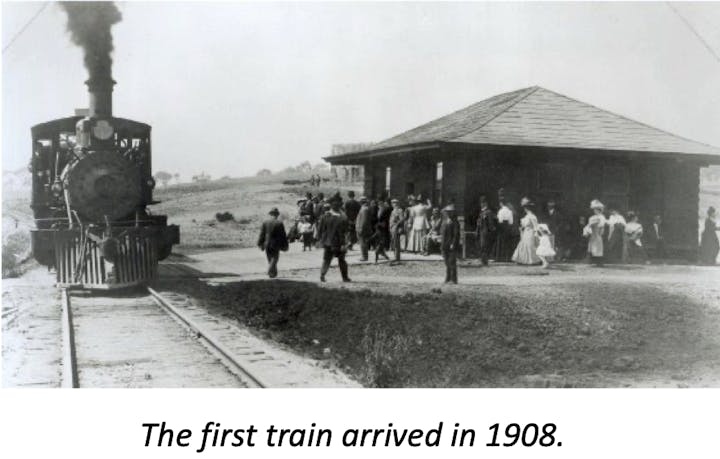 A steam train arrives at a station in 1908, with people gathered around.