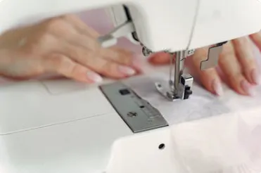 A close-up of hands using a sewing machine, with fabric being guided under the needle for stitching.