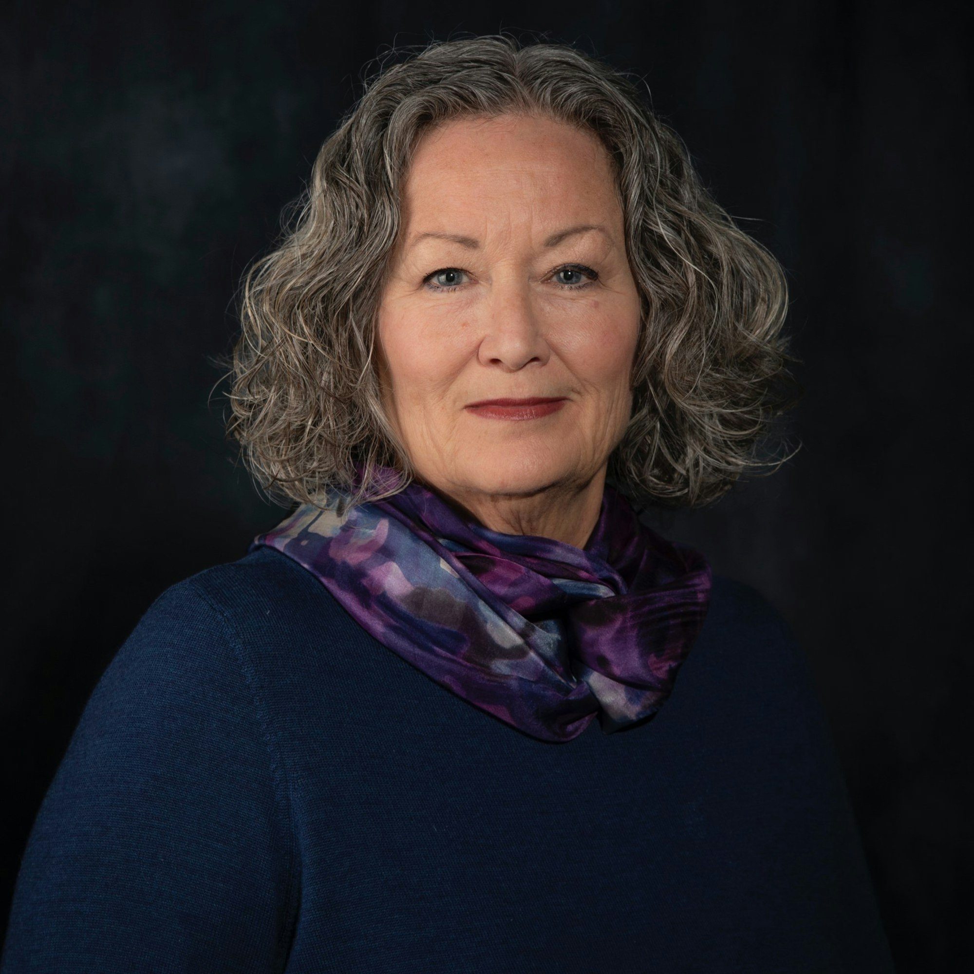 A person with curly gray hair wearing a dark blue top and a purple scarf, posing against a dark background.