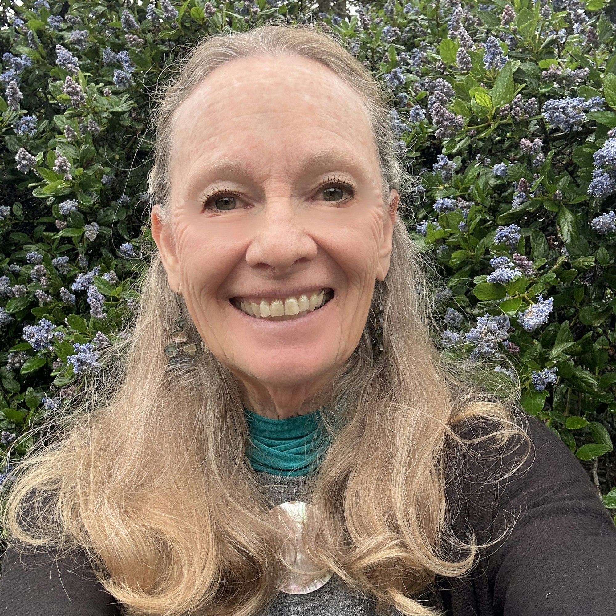 A person with long hair smiling in front of a bush with small purple flowers.