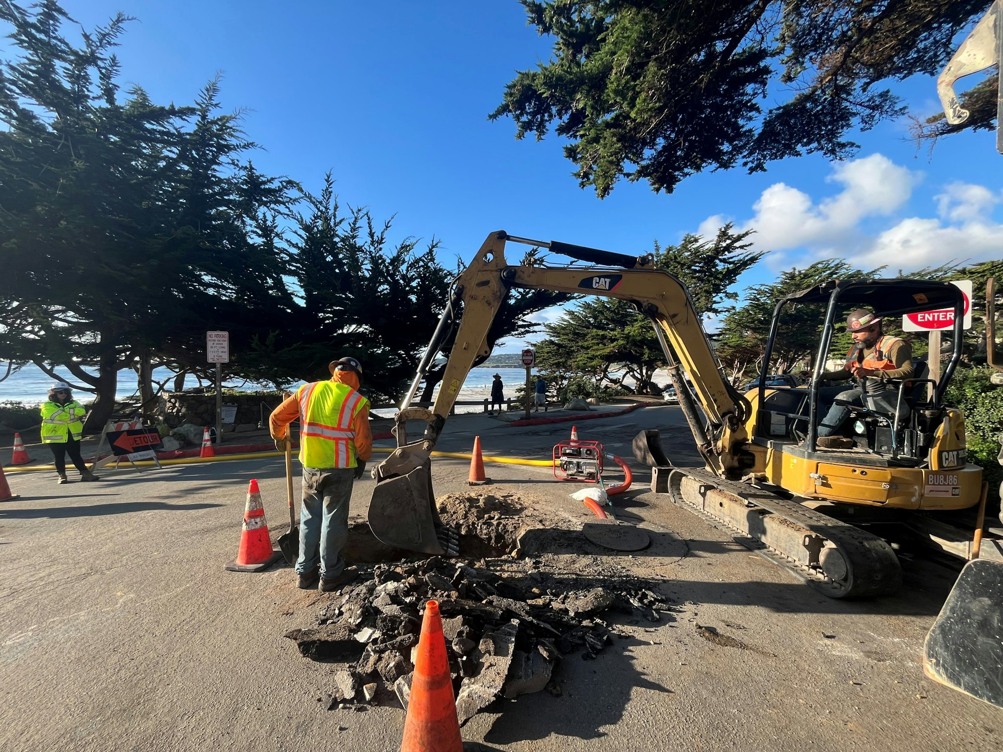 Construction workers with an excavator repairing a road, surrounded by traffic cones and trees.