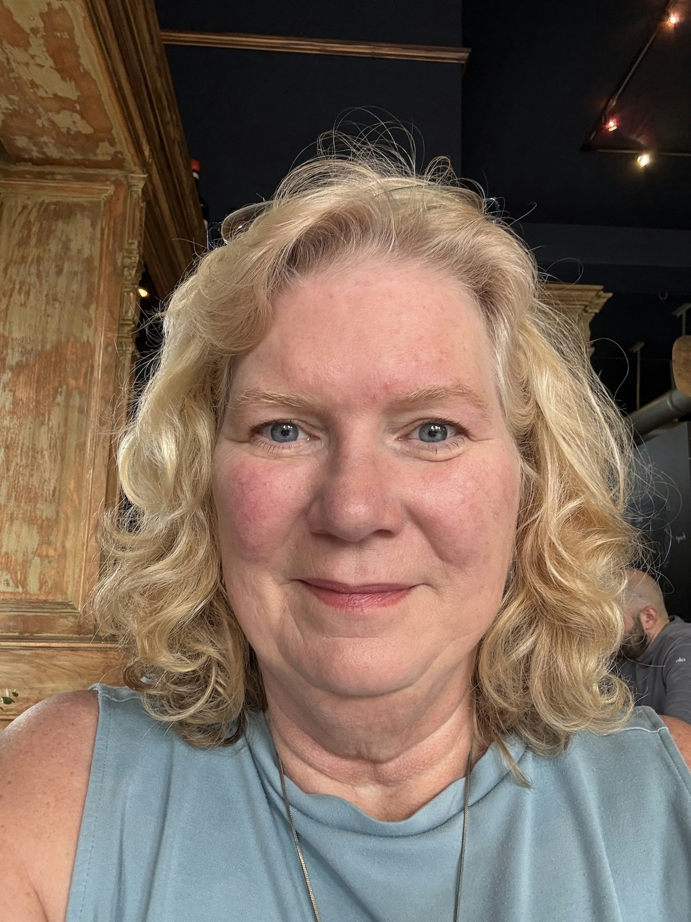 A person with curly blonde hair, wearing a blue top, in an indoor setting with wooden decor.