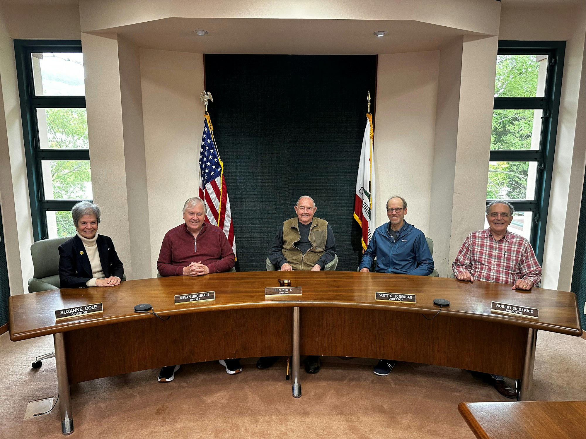 Five people sitting at a curved desk with nameplates, American flags in the background.