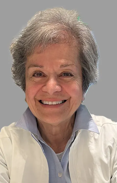 Smiling older person with short gray hair wearing a light jacket and collared shirt against a plain background.