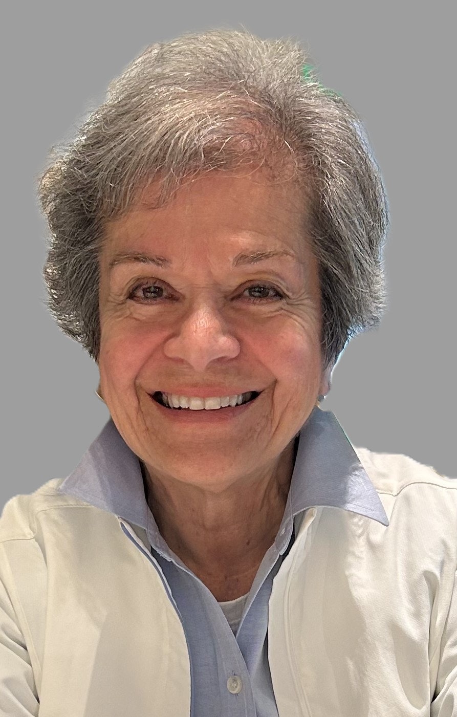 Smiling older person with short gray hair wearing a light jacket and collared shirt against a plain background.