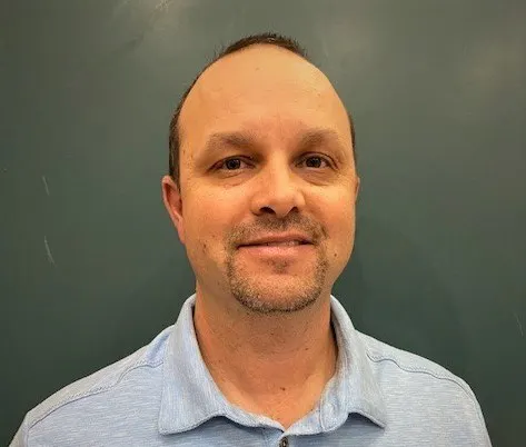 A smiling man with short hair, wearing a light blue shirt, stands in front of a green background.