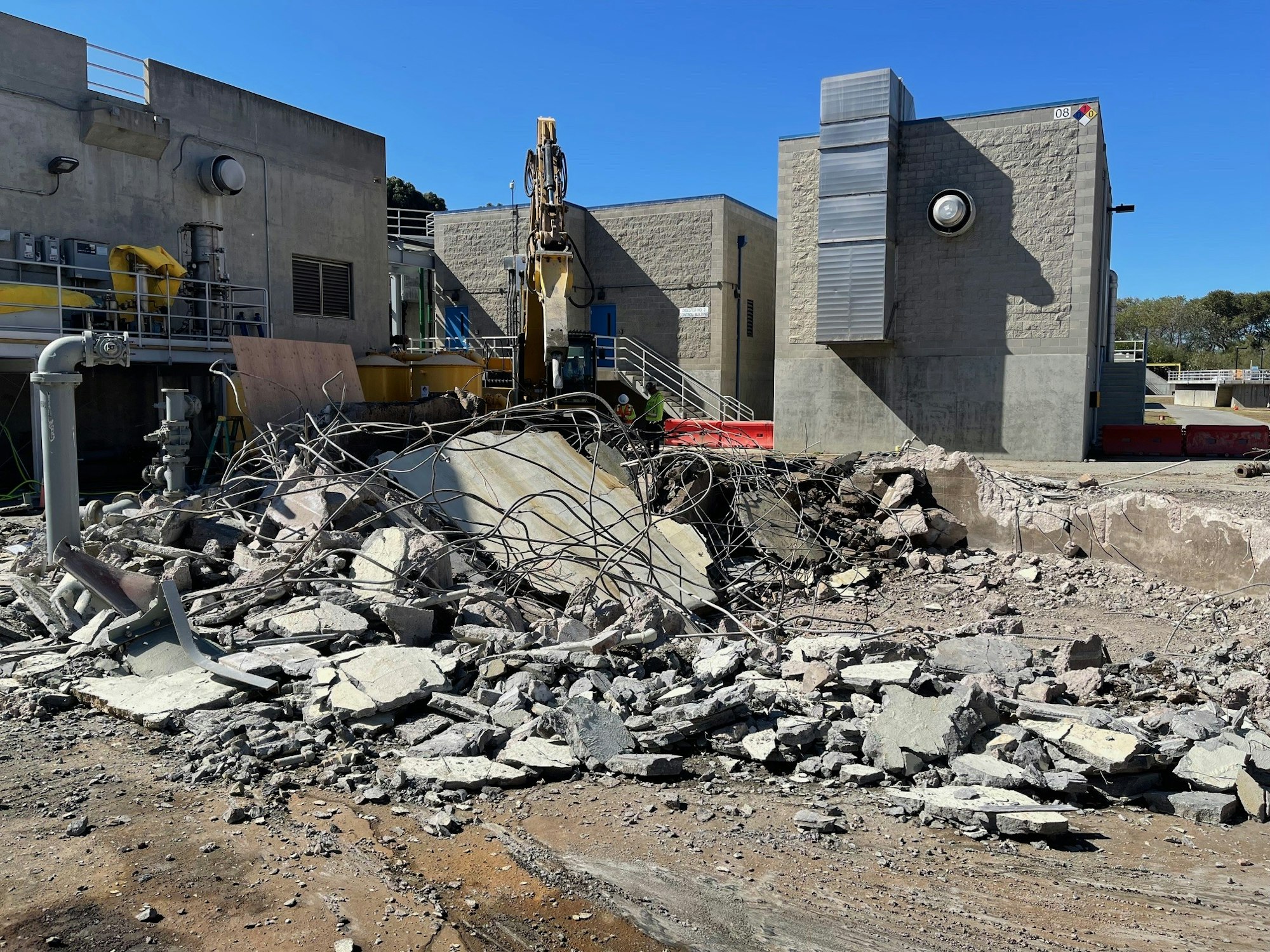 An industrial site with heavy machinery amidst piles of rubble and debris from concrete and steel structures.