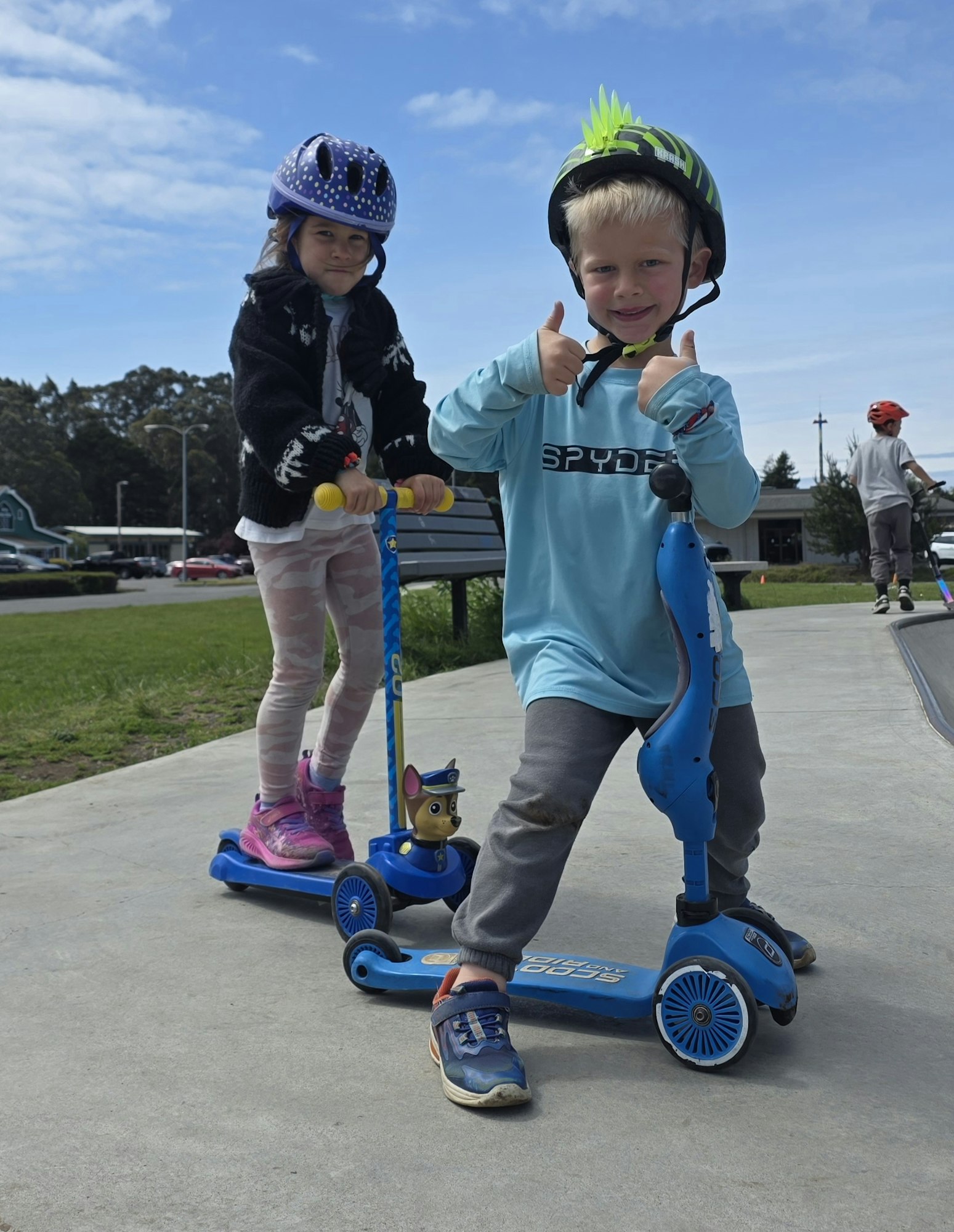 Two kids riding scooters at a park, wearing helmets and enjoying a sunny day. The boy gives a thumbs up.