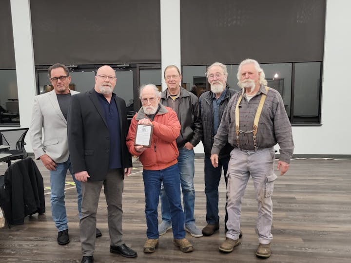 Six men standing indoors; one holds a plaque. Casual to semi-formal attire.