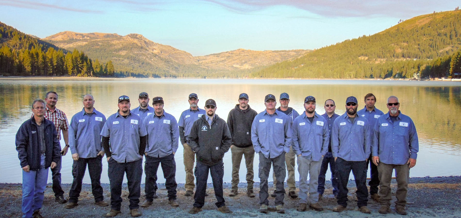 A group of men in matching shirts and hats pose together by a lake, surrounded by trees and mountains.