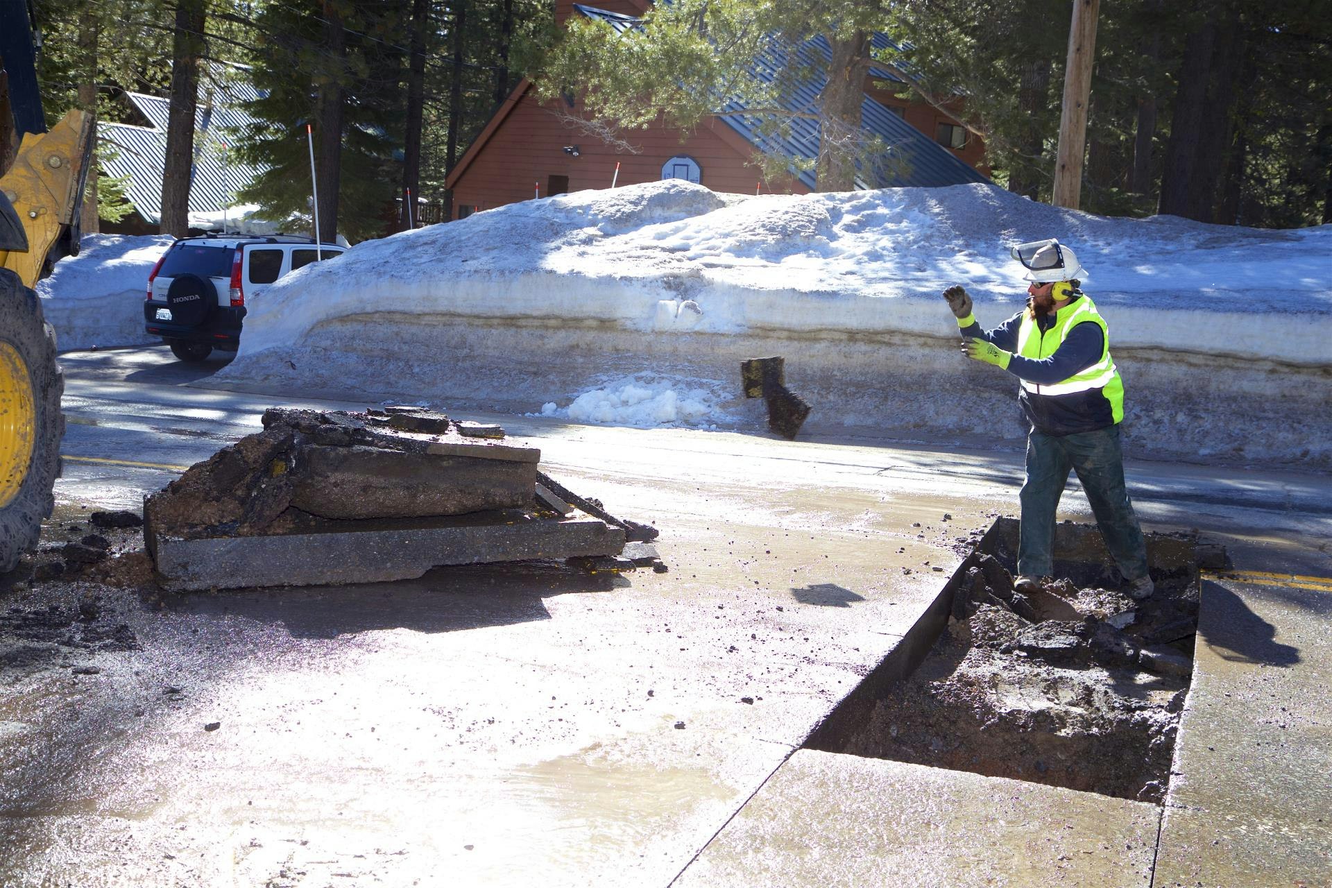 A construction worker is repairing a damaged road, with snow in the background and a vehicle parked nearby.