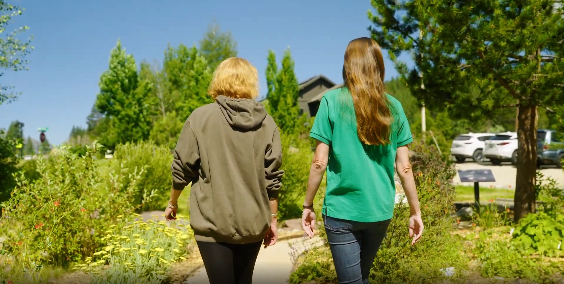Two women walk through a lush garden on a sunny day, surrounded by greenery and parked cars in the background.