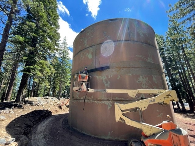A large, rusted metal tank is being serviced by a worker using a lift in a forested area with trees and blue skies.