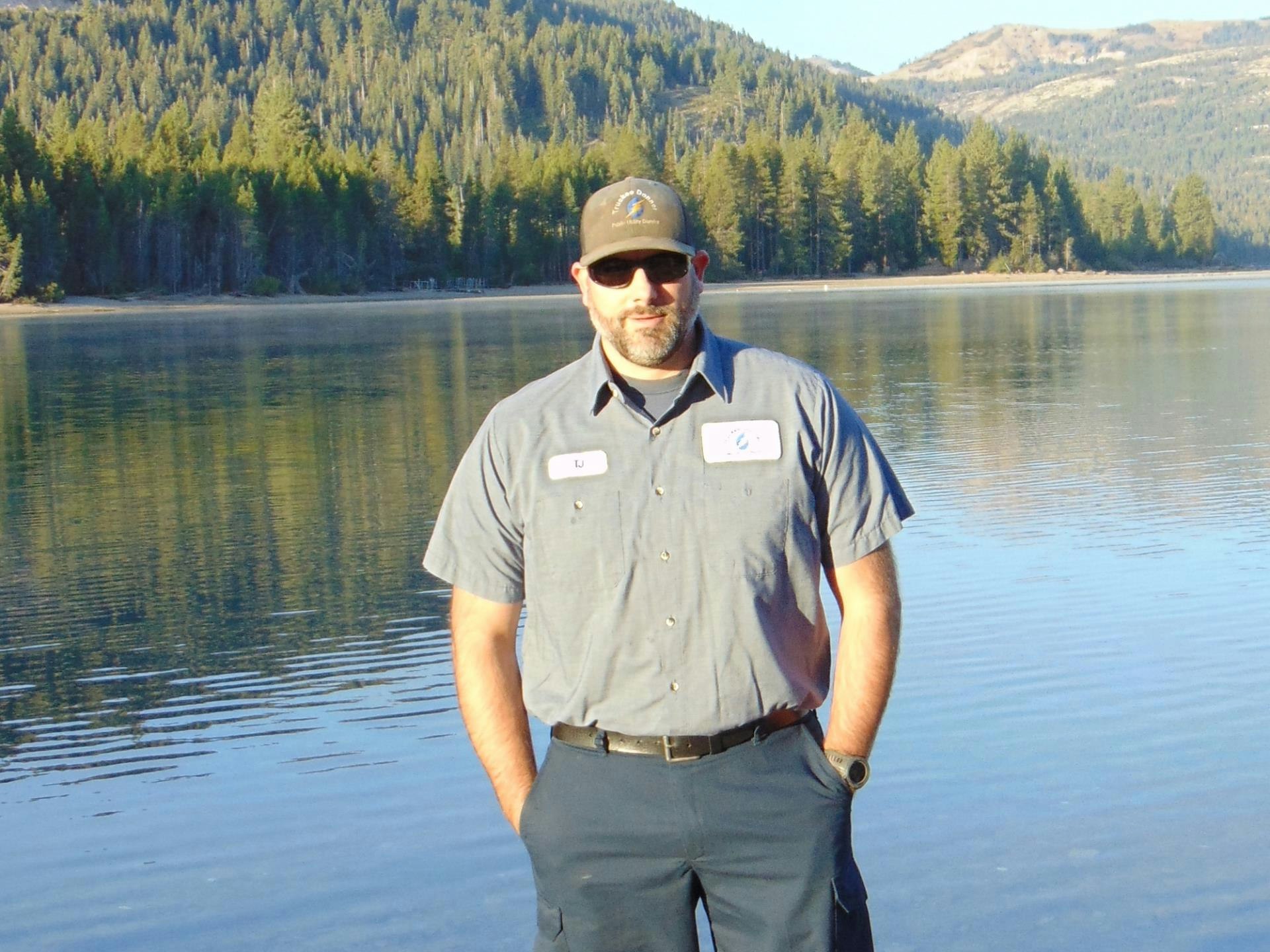 A person stands by a lake surrounded by trees, wearing a shirt with patches and a cap. The scene is calm and scenic.