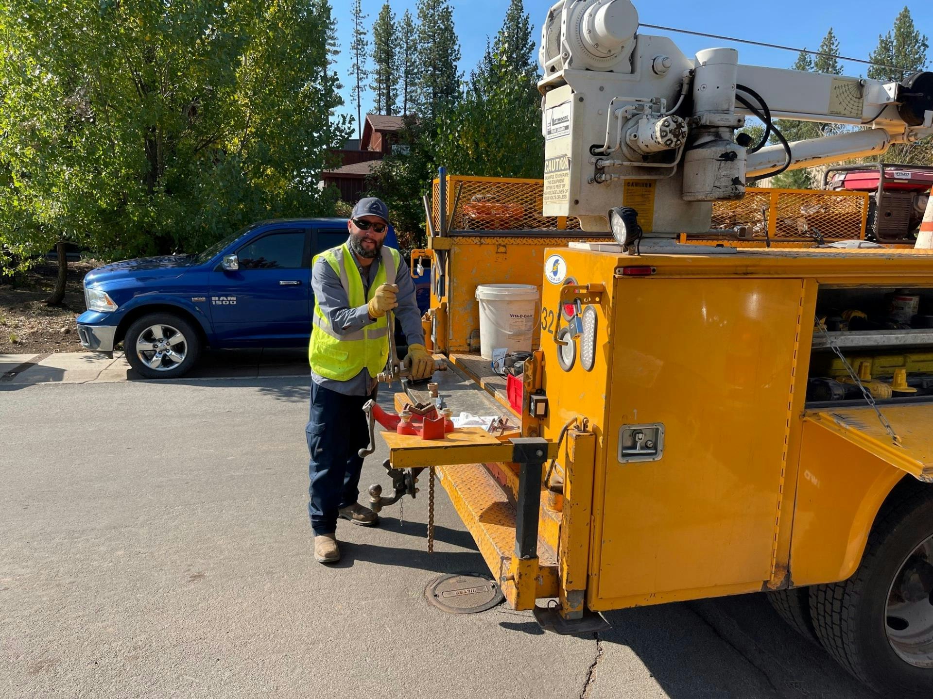 A man in work gear stands by a yellow utility truck, preparing tools for work. A blue pickup is visible in the background.
