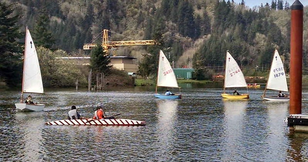 Sailboats on the water, one boat with striped design, forest and crane in the background.