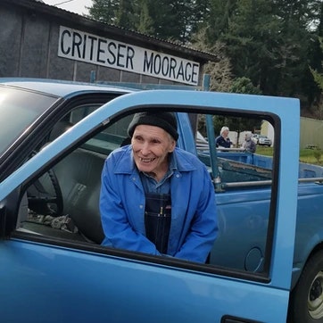 An elderly person smiling from the driver's seat of a blue pickup truck, with a "CRITESER MOORAGE" sign in the background.