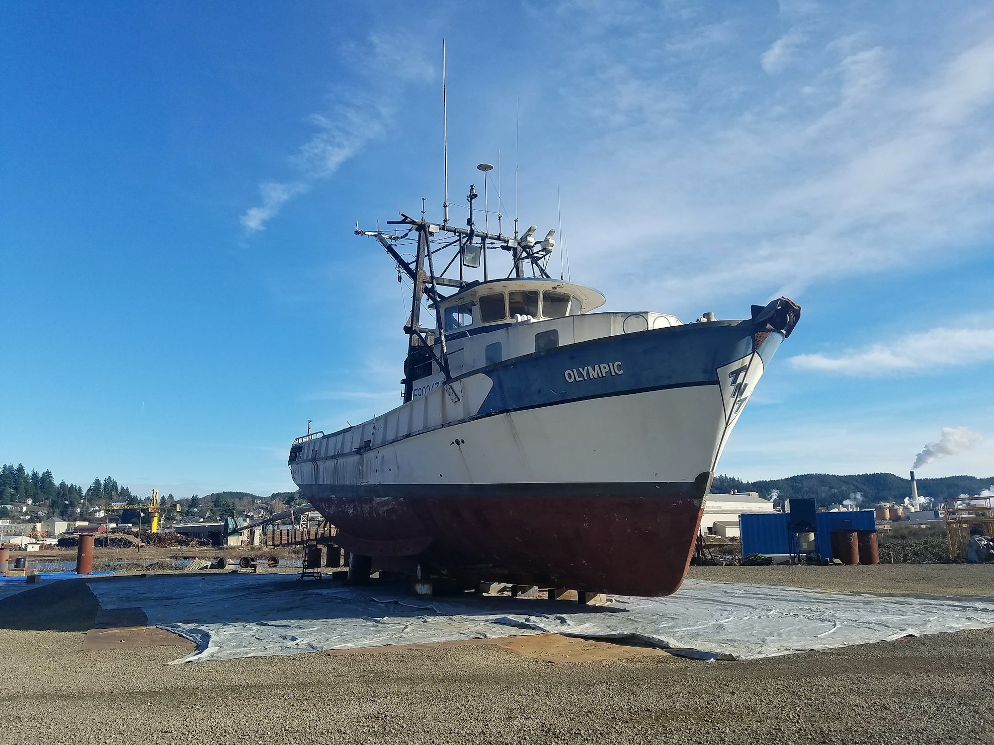 A fishing boat named "OLYMPIC" is dry-docked on land under a clear blue sky.