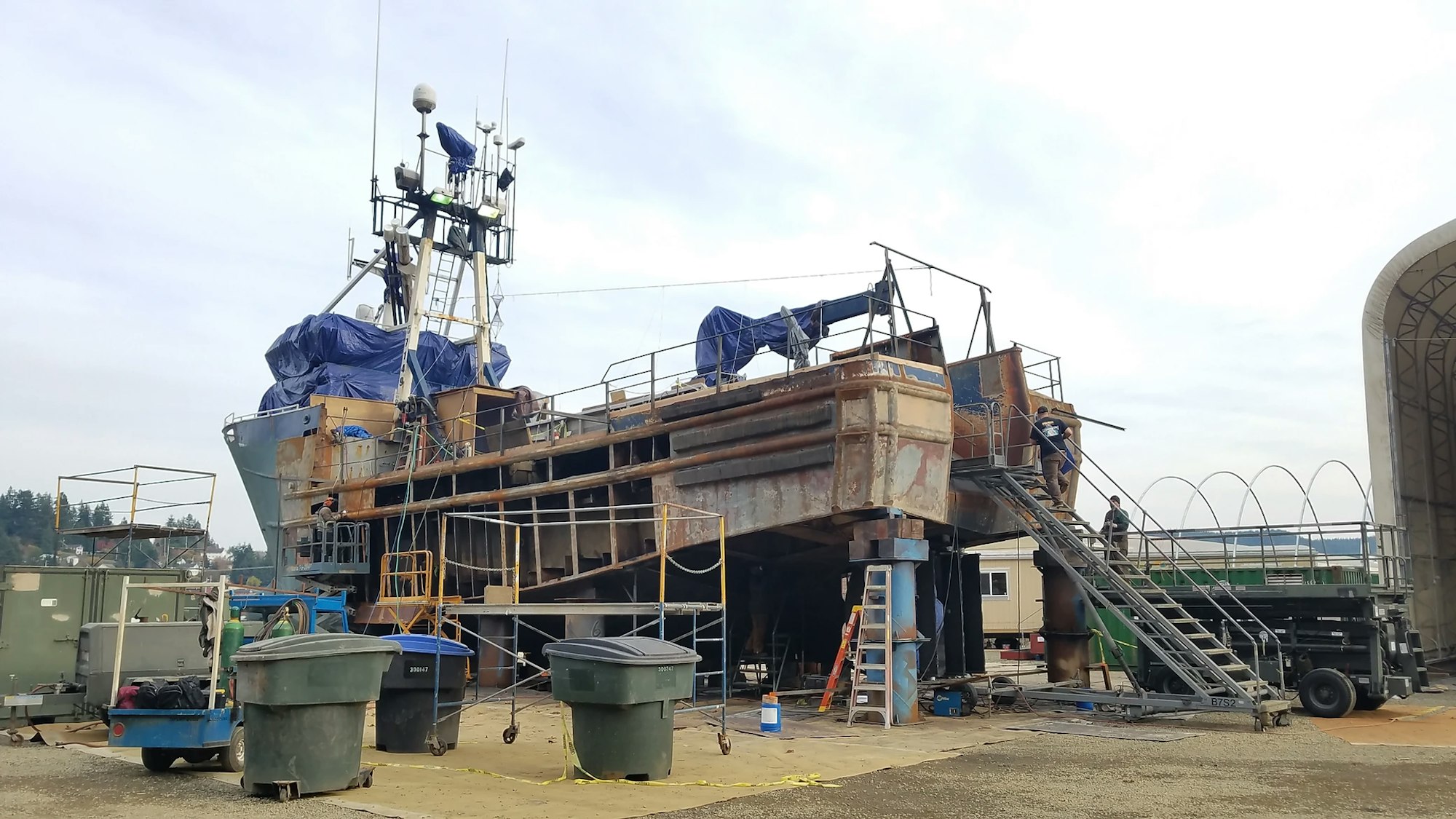 A large boat undergoing repairs or maintenance at a shipyard with equipment and workers.