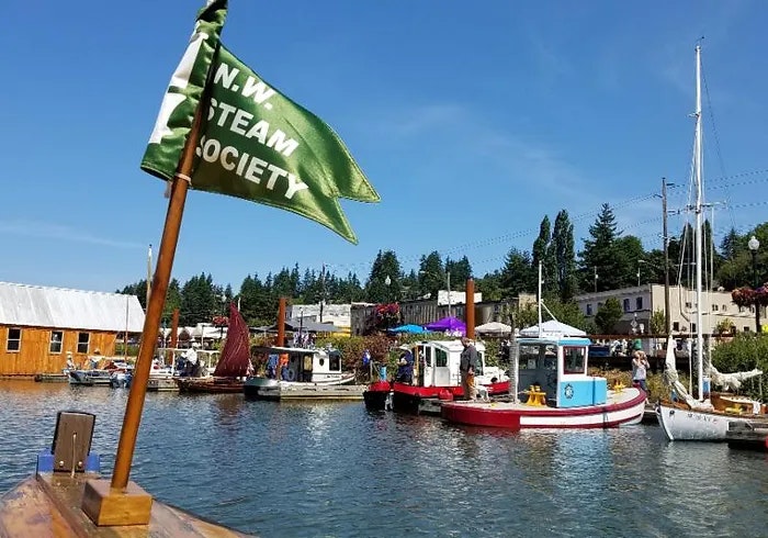 A flag reading "N.W. STEAM SOCIETY" with boats on water, clear blue sky, and buildings in the background.