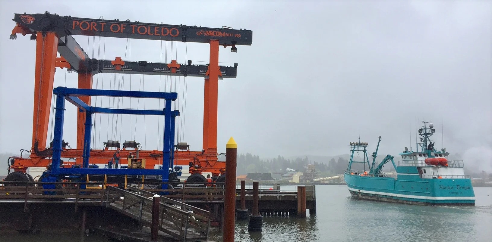 A ship named Alaska Trojan docked at Port of Toledo with a large orange marine travelift on a misty day.
