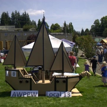 Cardboard boat display at an outdoor event with people in the background.