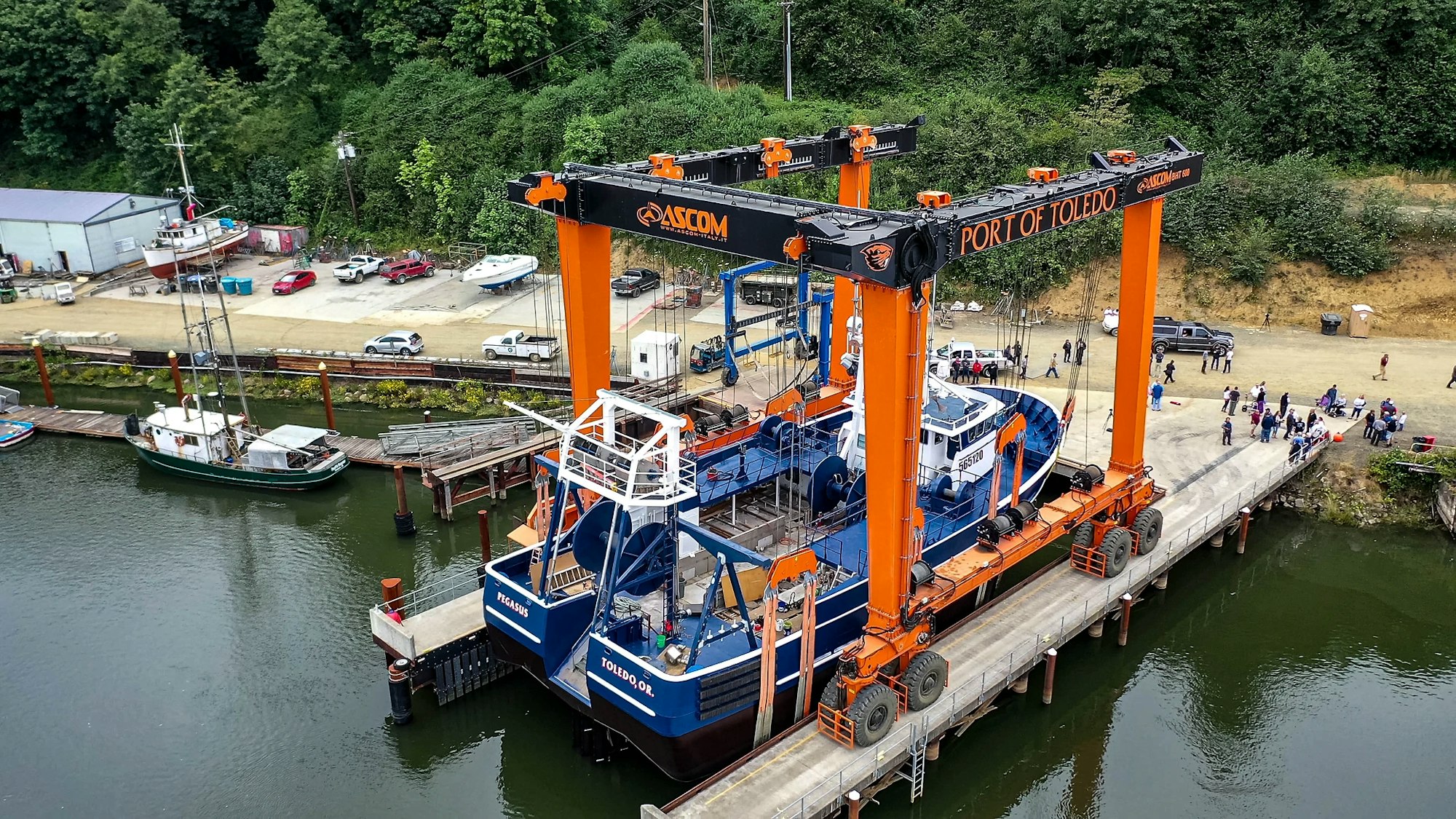 Aerial view of a marine industrial scene with a large orange boat lift at the Port of Toledo, surrounded by boats, vehicles, and people.