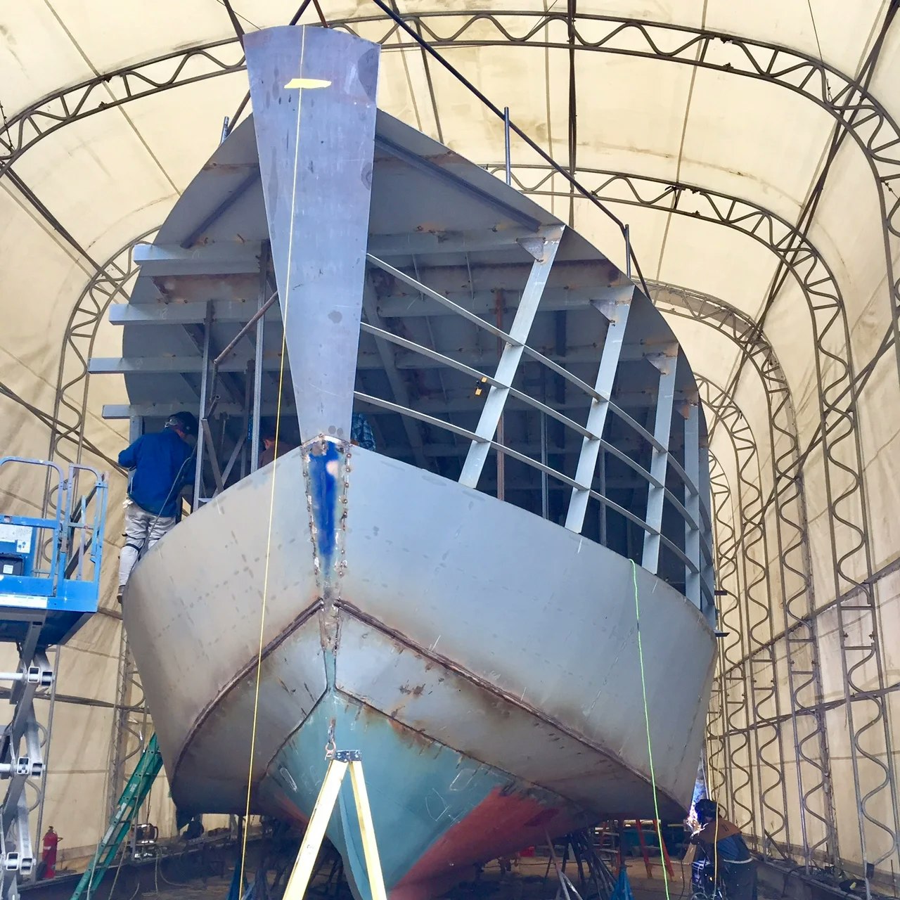 A ship under construction or repair in a sheltered workspace with workers present.
