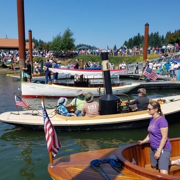 A gathering by a body of water with people on boats displaying American flags; a crowd observes from the shore.