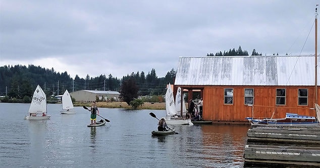 Sailboats on water near a rustic building, with individuals on a dock and paddleboarding. Cloudy sky overhead.