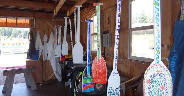 Colorful paddles and life jackets inside a wooden shed, possibly at a lakeside or beach.
