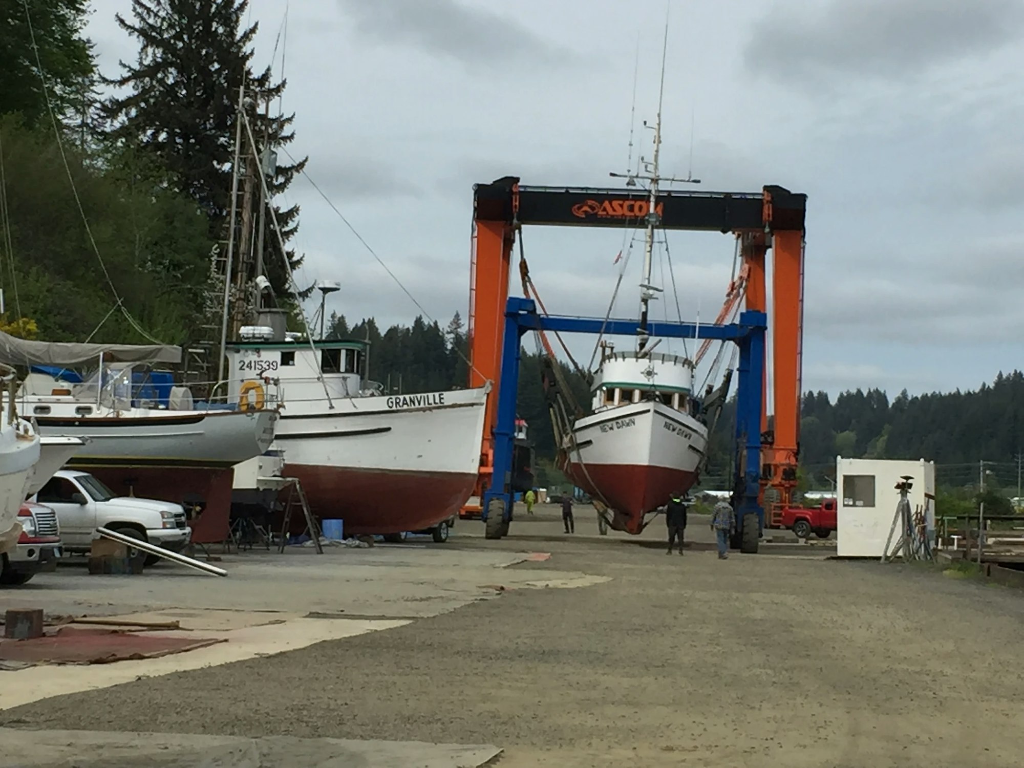 A boatyard with a large blue and orange boat hoist lifting a fishing vessel, with people and other boats around.