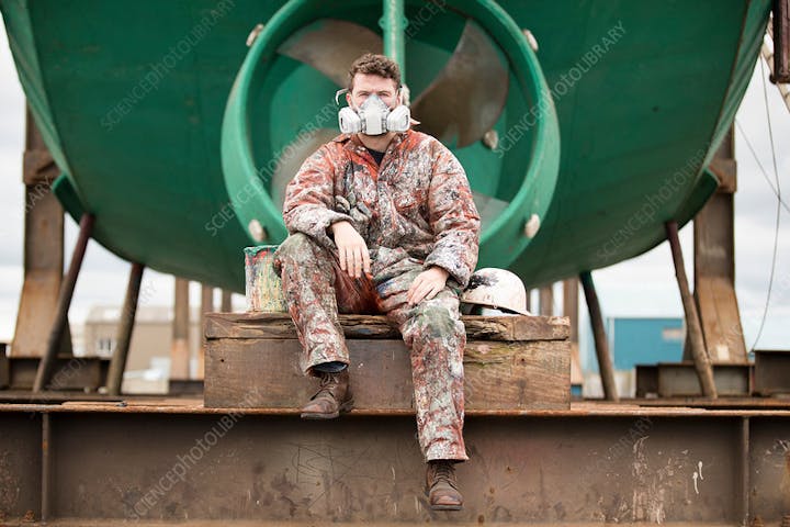 Worker in paint-splattered overalls and mask sitting on platform, with a large industrial object in the background.