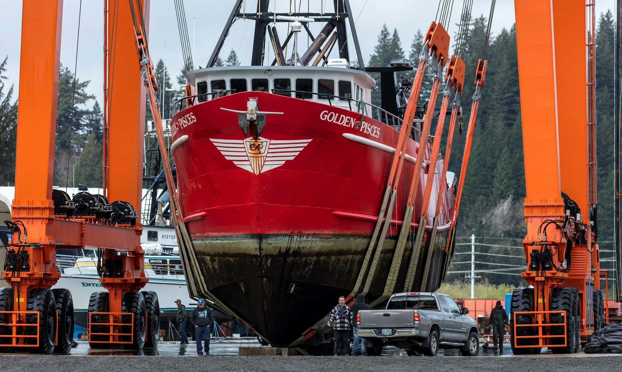 Red fishing vessel "Golden Pisces" held by a large orange mobile boat hoist with people and a truck nearby.