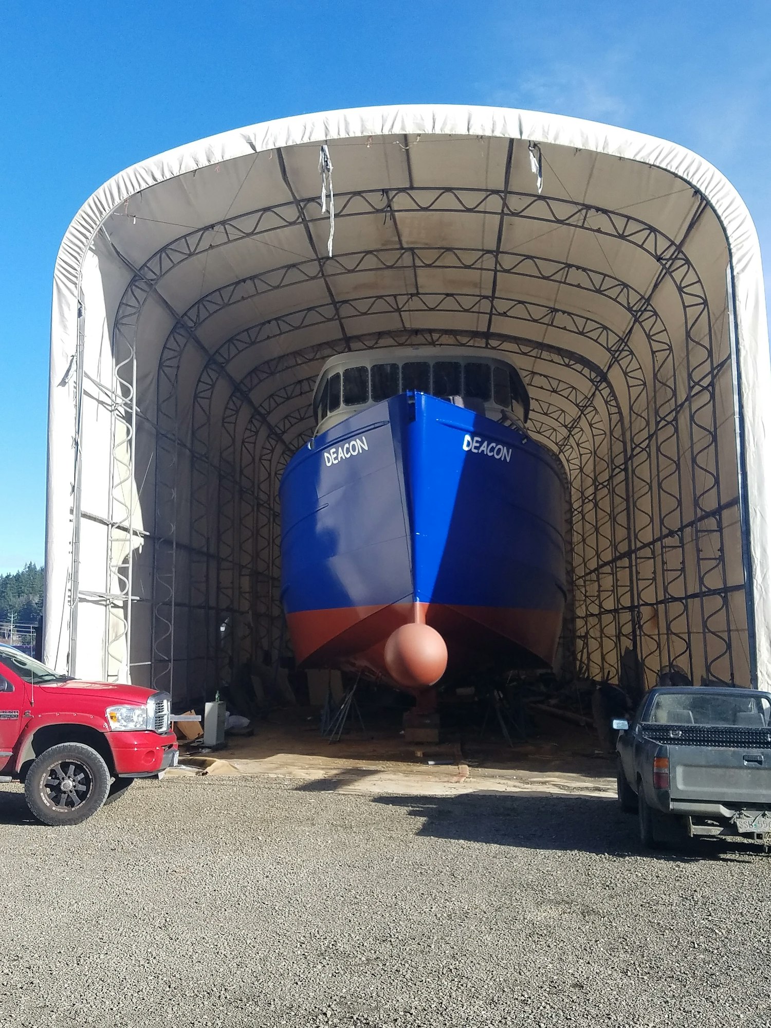 Large ship in a dry dock tent, blue and orange hull, clear sky, trucks nearby.