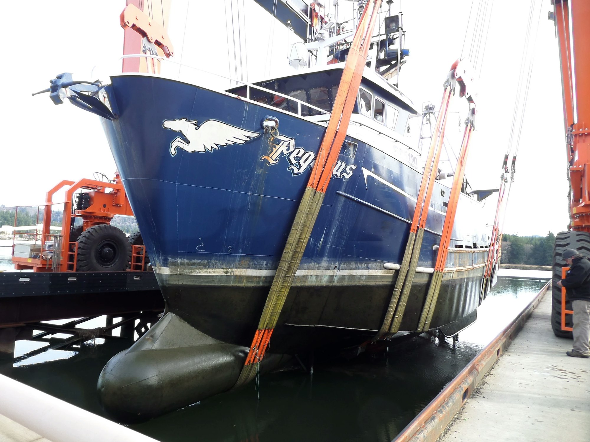 A blue fishing vessel named "Kiska Sea" in a dry dock, supported by straps and blocks, with a forklift nearby.
