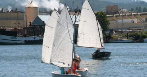 Two sailboats on water with industrial buildings in the background.