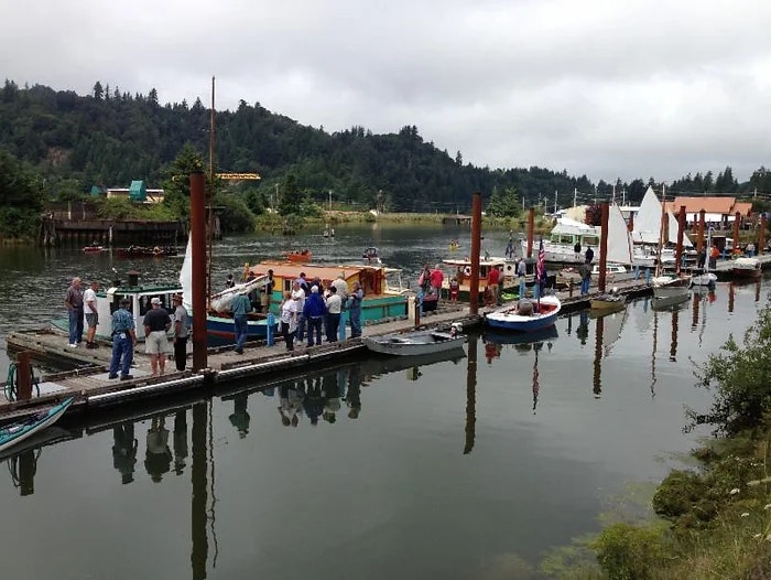 A wooden dock with people and moored boats, set against a backdrop of hills and overcast sky.