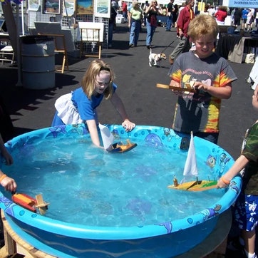 Children play with toy boats in a small inflatable pool at an outdoor event.