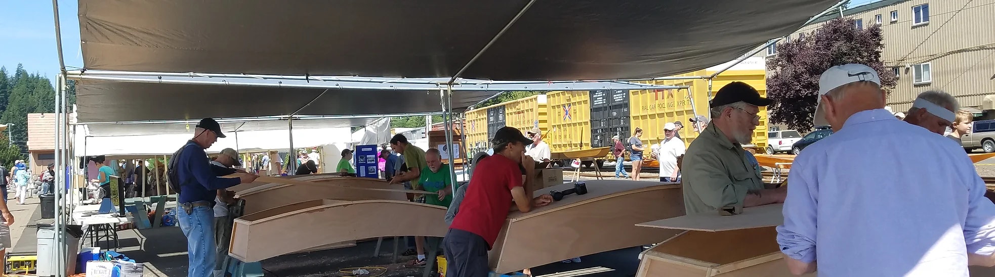 Outdoor workshop with people building wooden boats under a tent, surrounded by trees and clear blue sky.