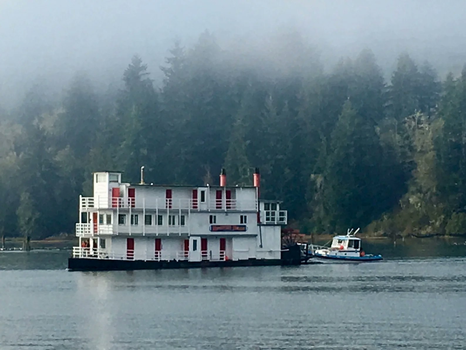 A riverboat on a misty river with a tugboat and forested hills in the background.