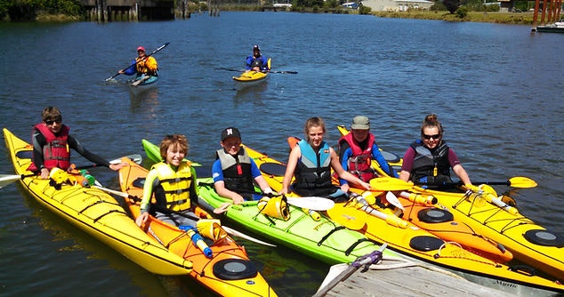 Group of people in life jackets on yellow kayaks with paddles, sunny day on calm water.