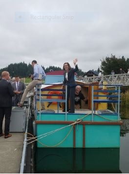 People on a boat with one person waving, moored at a dock with a cloudy sky.