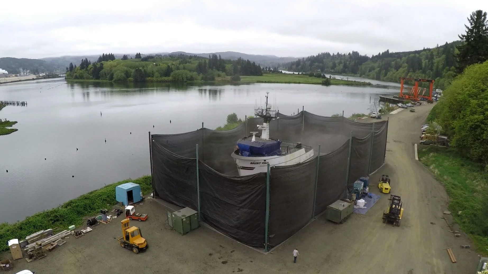 Aerial view of a boat encased in netting near a river with equipment and vehicles onshore.