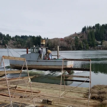 A boat near a dock with a person on board, surrounded by calm water and greenery in the background.
