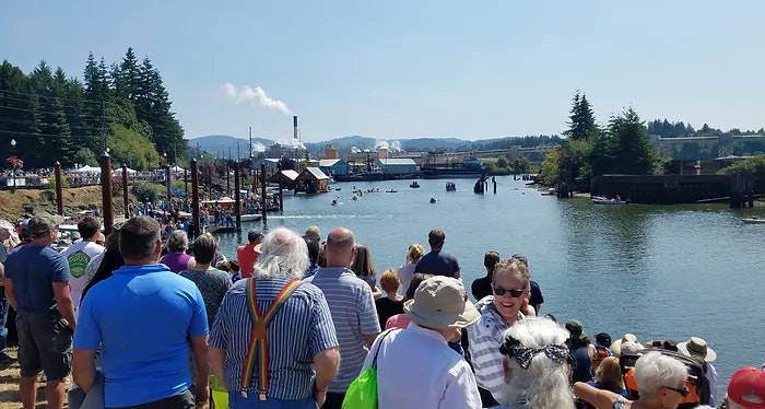 Crowd of people by a waterway on a sunny day, with industrial buildings in the distance.