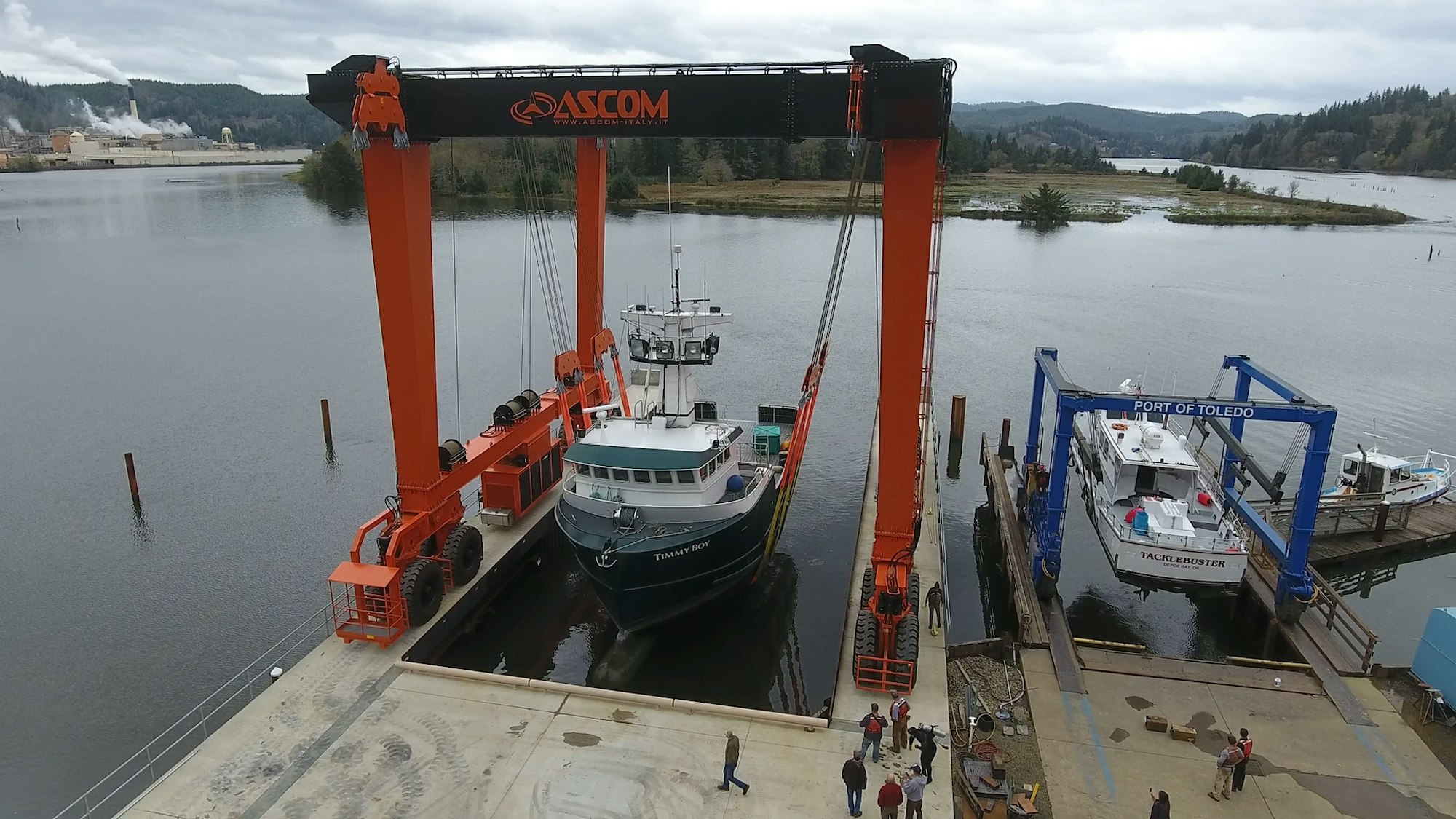 A shipyard with boats, a travel lift, and people, by a river with industrial structures in the background.