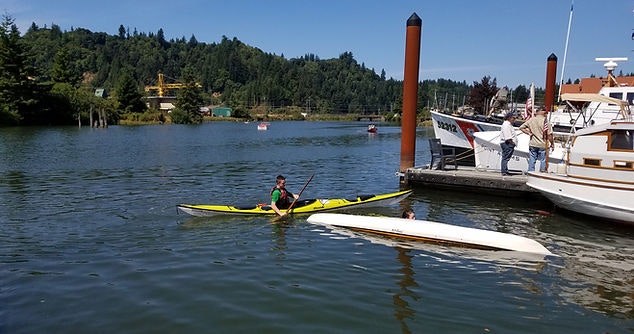 A person kayaking near a dock with boats on a sunny day, with a wooded area in the background.