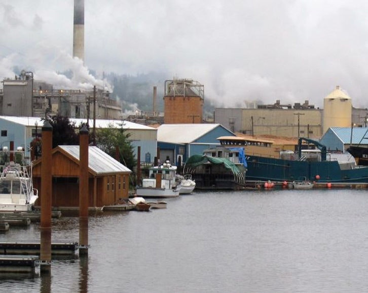 Industrial factory by the water with smokestacks and boats docked nearby. Cloudy sky.
