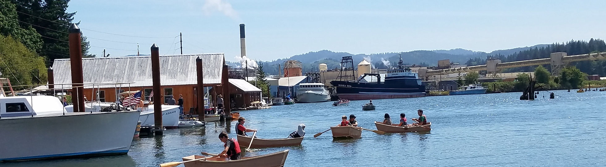 People rowing boats in a harbor with industrial buildings in the background.
